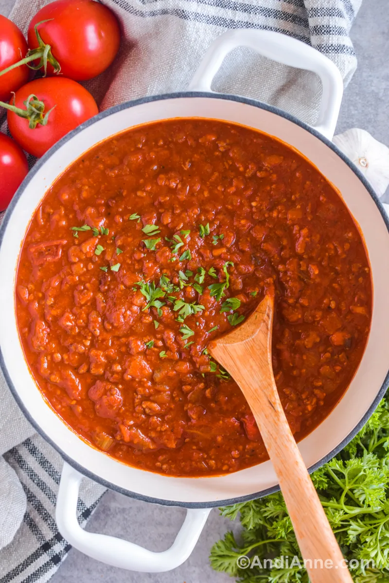 A large white pot of ground beef spaghetti sauce with chopped parsley sprinkled overtop. Tomatoes, a kitchen towel and fresh parsley surround the pot.