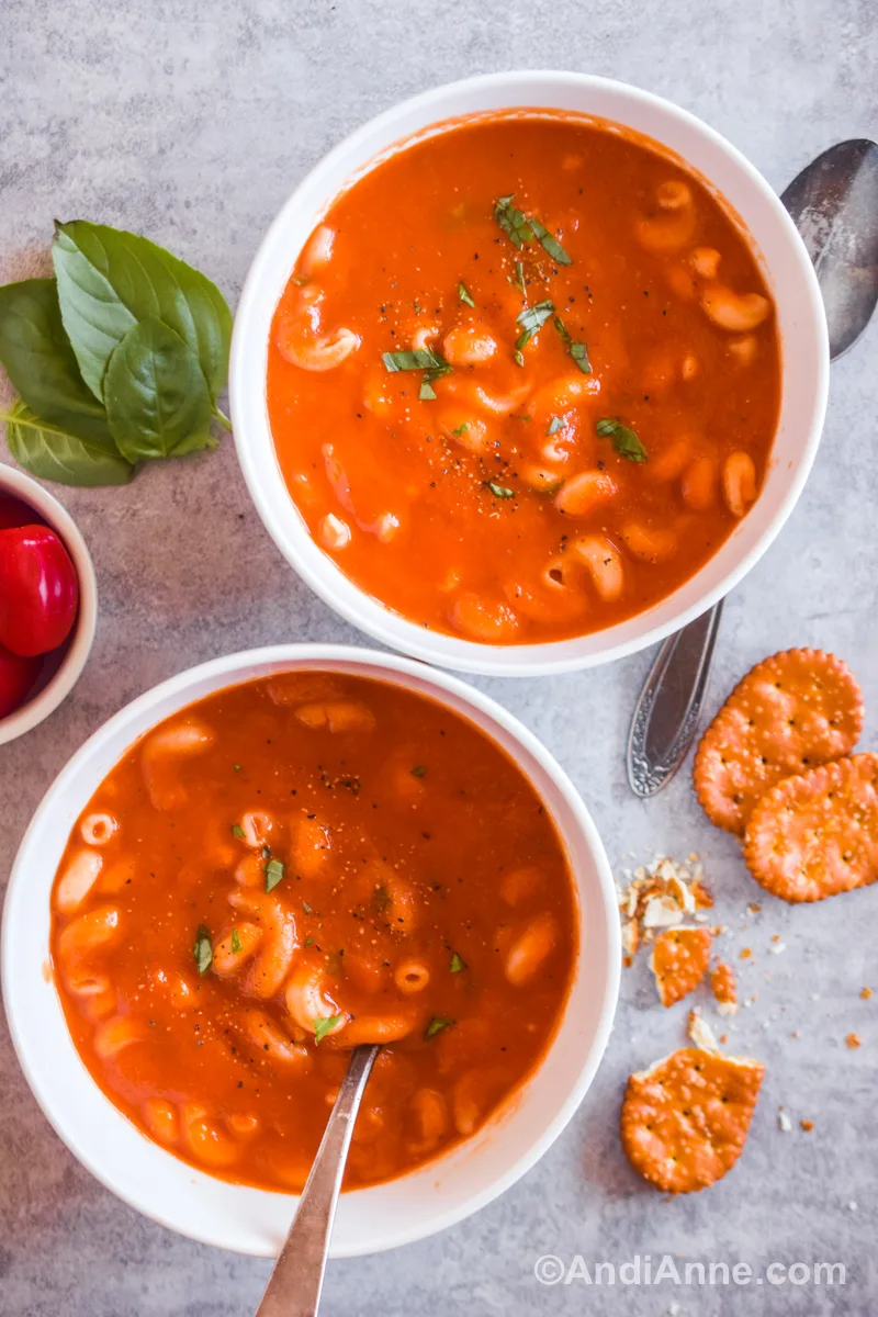 Two bowls of tomato pasta soup seasoned with chopped basil, salt and pepper. Spoons, a few crackers, grape tomatoes and some basil leaves surround the bowls.