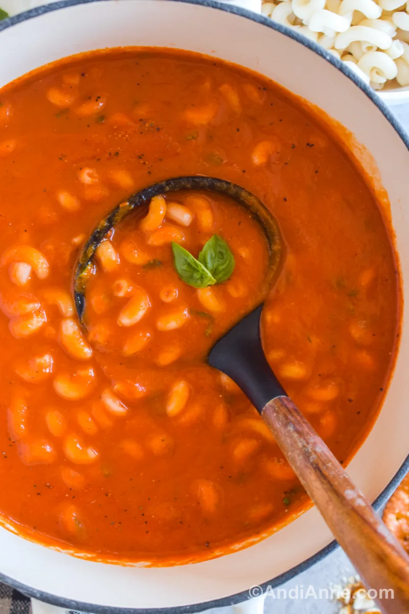 A pot of tomato pasta soup with a soup ladle and two basil leaves.