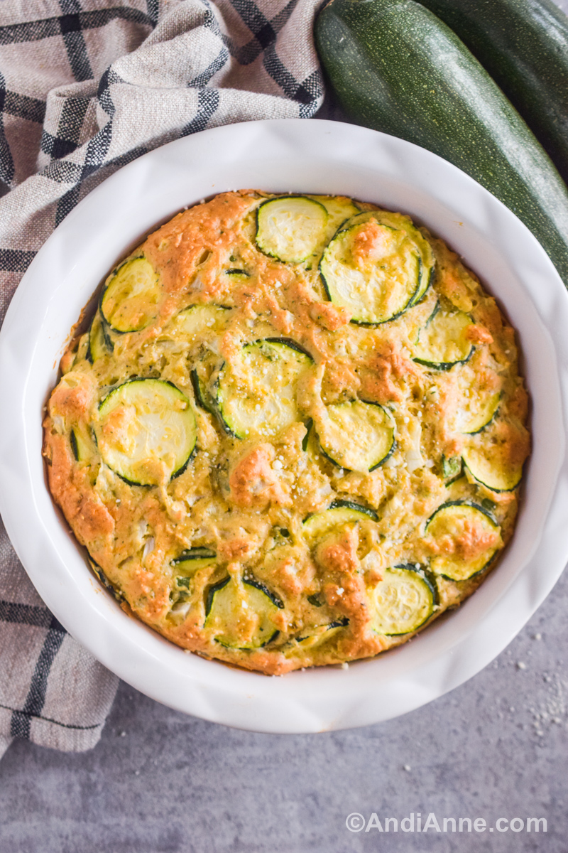 Zucchini pie in a pie plate with fresh zucchinis and a kitchen towel surrounding the plate.