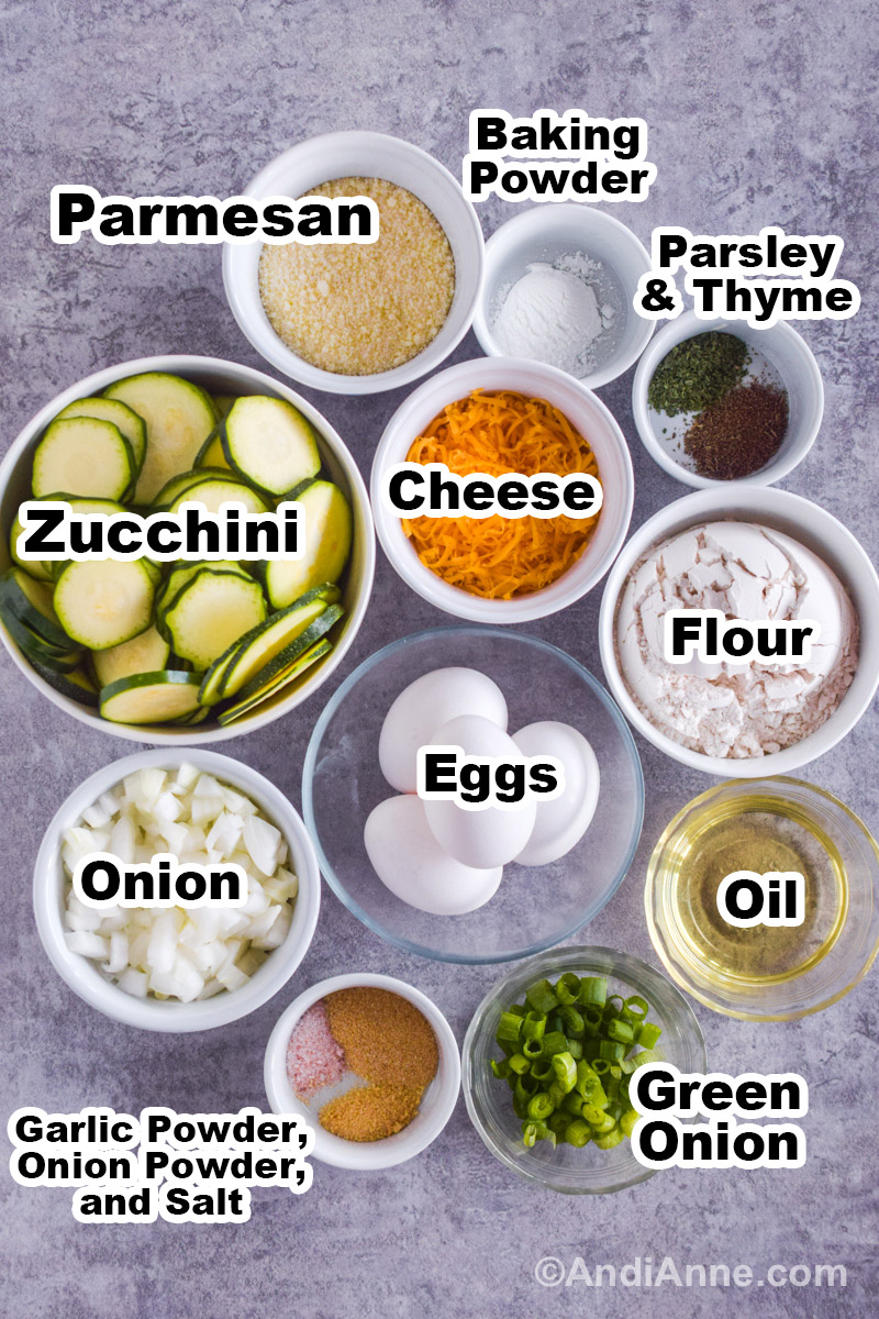 Recipe ingredients on a counter including bowls of sliced zucchini, chopped onion, green onions, parmesan cheese, grated cheddar cheese, baking powder, parsley, thyme, garlic powder, onion powder and salt.