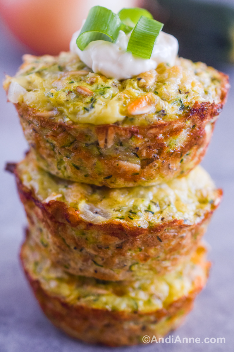 Three parmesan zucchini bites stacked on top of each other with a dollop of sour cream and three chopped green onions.