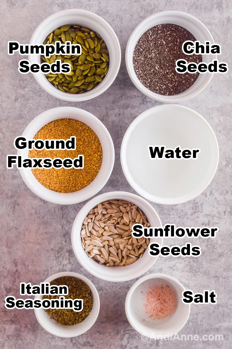 Recipe ingredients on a counter including bowls of pumpkin seeds, chia seeds, ground flaxseed, water, sunflower seeds, italian seasoning and salt.