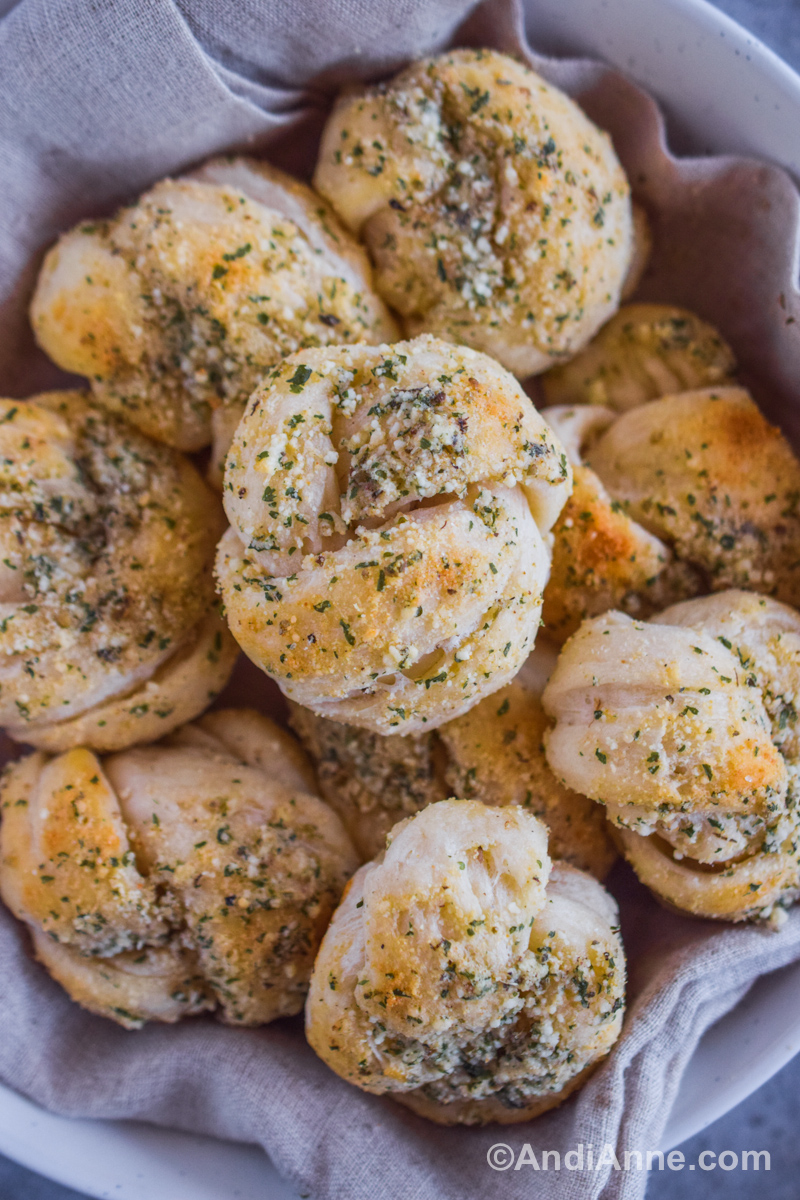Piled garlic parmesan knots in a basket with a kitchen towel underneath.