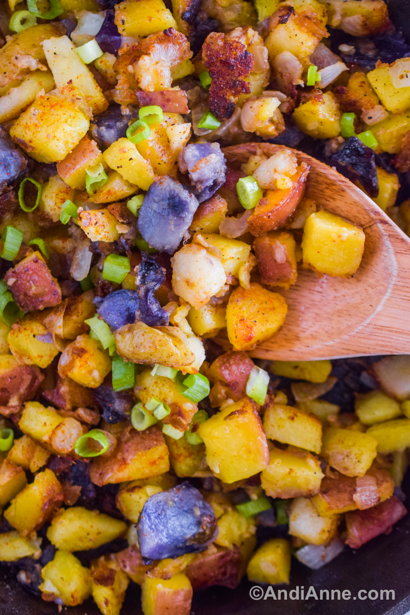 Close up of chopped yellow, purple and red potatoes with green onions and a wood spoon.