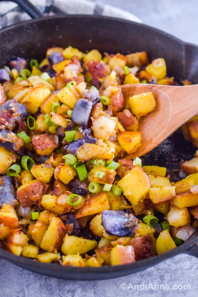 close up of chopped potatoes, green onions and spices in hash browns in a skillet with a wood spoon.