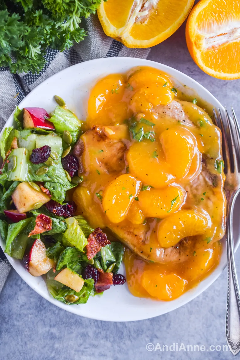 A cooked pork chop smothered in orange sauce with mandarin oranges and our fall apple salad beside it, all on a plate with a fork.
