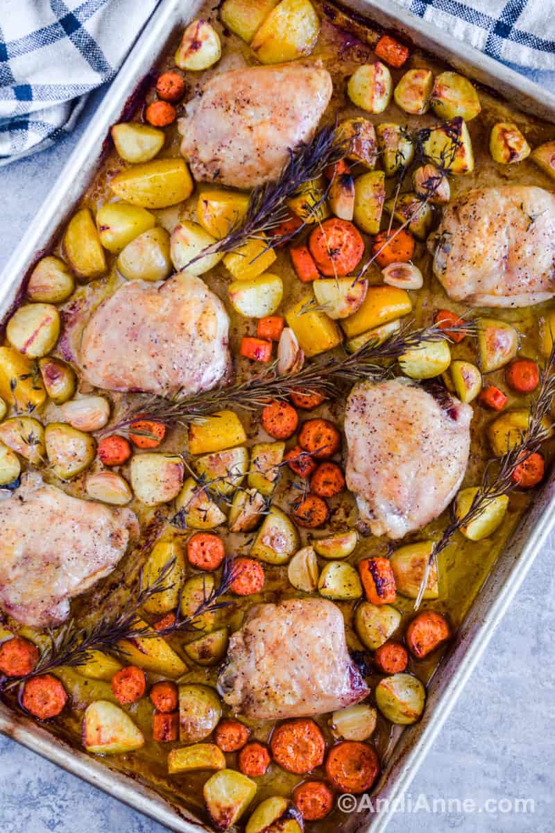 Looking down at a sheet pan with baked chicken thighs, chopped potatoes, and carrots. Cooked rosemary and thyme sprigs lay on top.