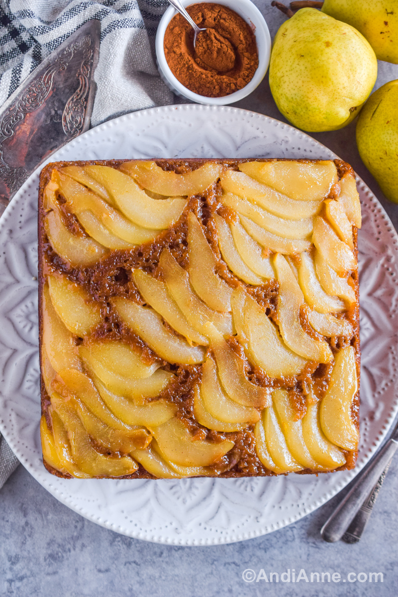 Upside down pear cake on a white dish with 3 pears, a bowl of cinnamon and a spoon, and a pie cutter on the side.
