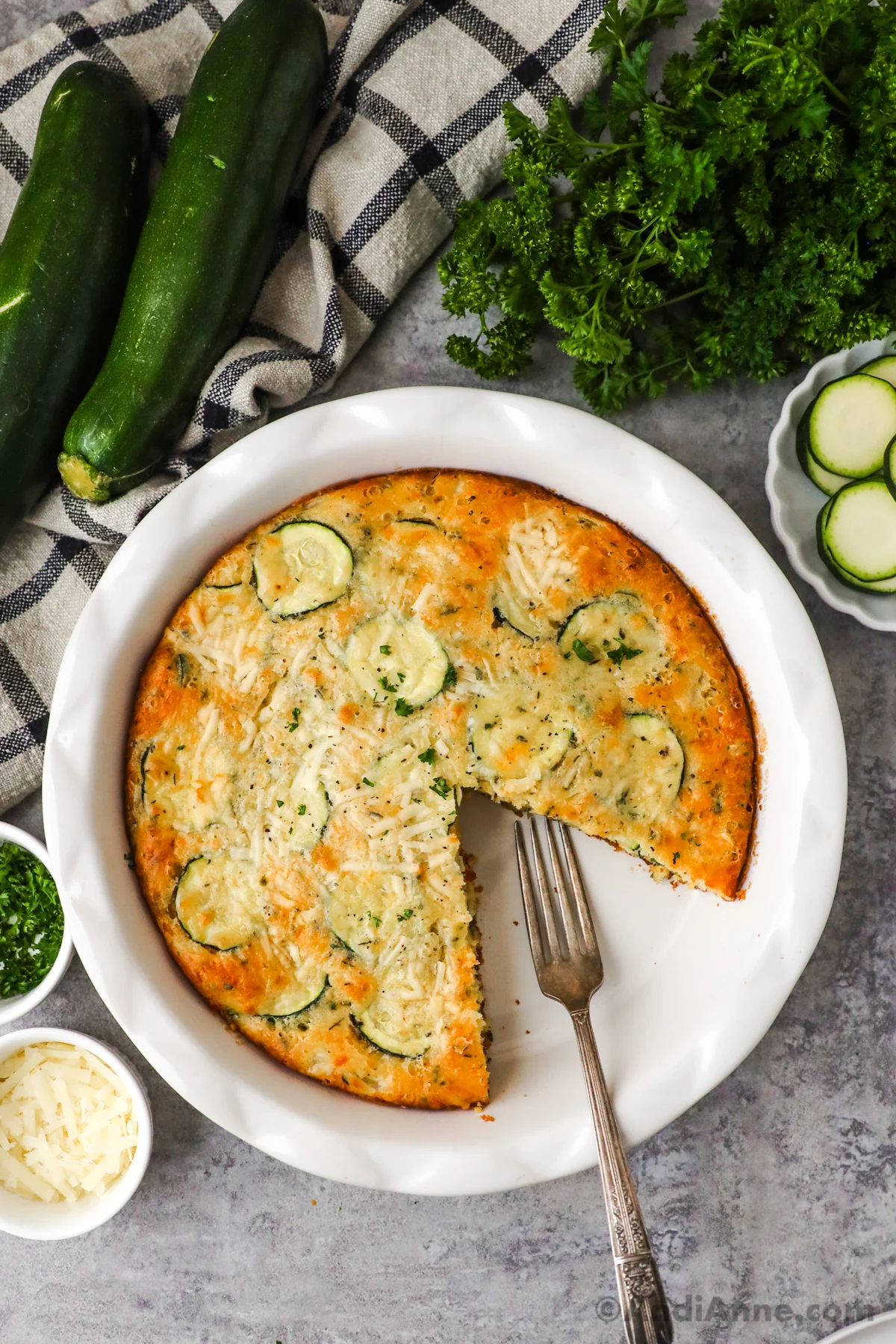 A slice cut out of zucchini pie with a fork, surrounded by bowls of ingredients, fresh zucchini and parmesan.