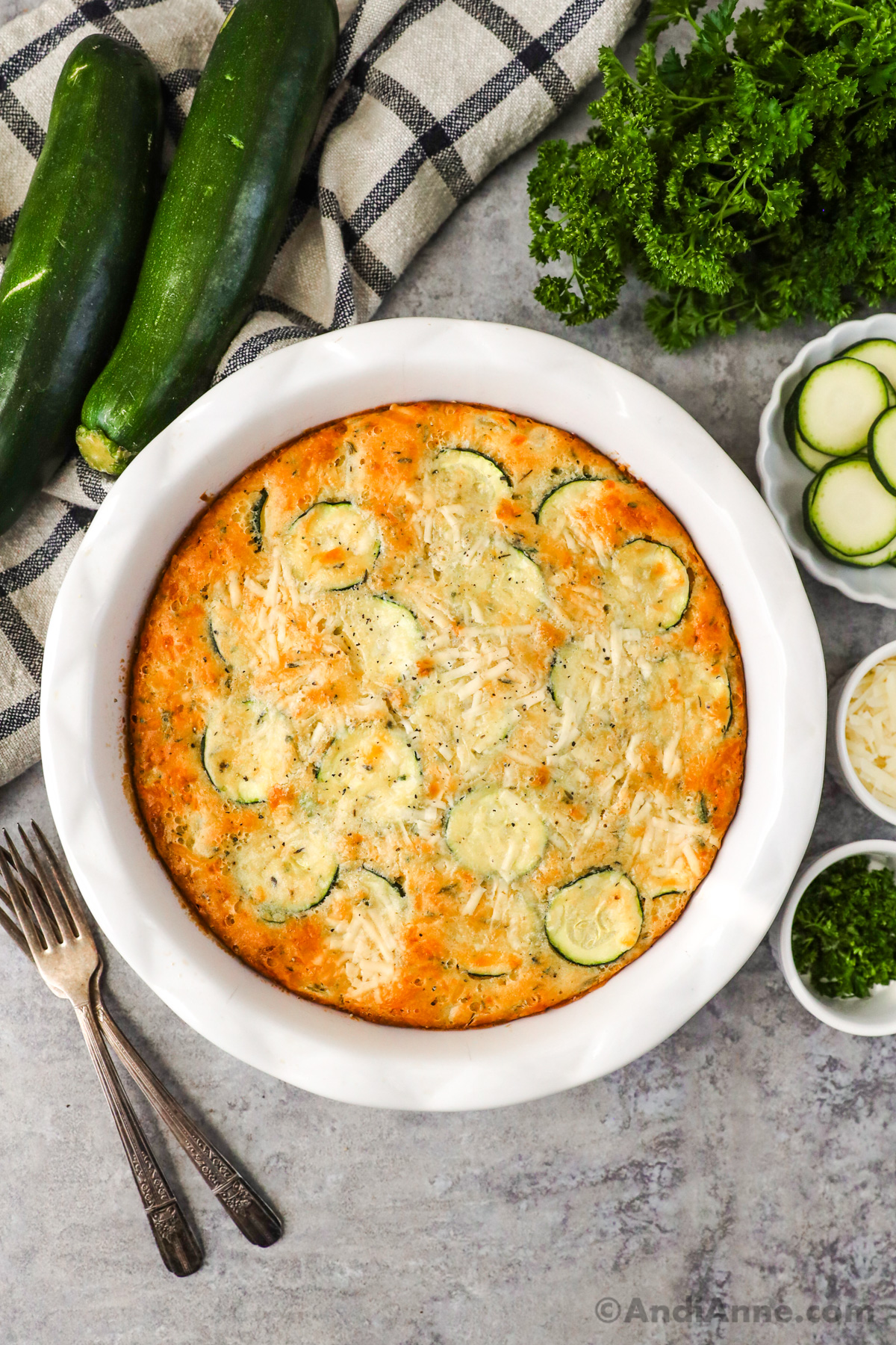 Baked zucchini pie in a white pie dish surrounded with forks, zucchini, bowls of shredded cheese, sliced zucchini and parmesan.