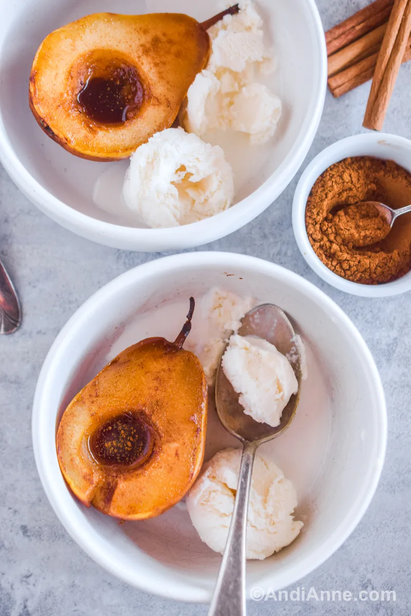 Two bowls with a slice of baked pear, and two scoops of ice cream and a spoon. A small bowl with cinnamon and cinnamon sticks beside them. 