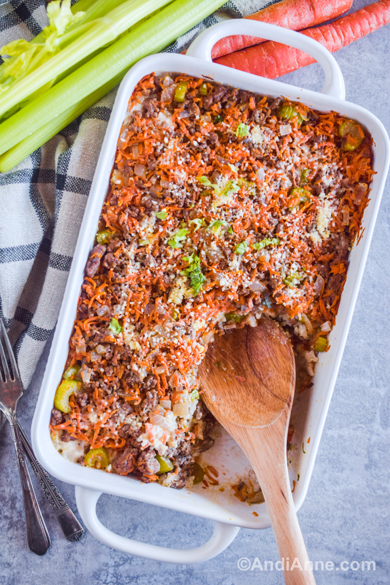 A white casserole dish with beef and rice casserole recipe inside plus a wood spoon. Forks, carrots and celery surround the dish.