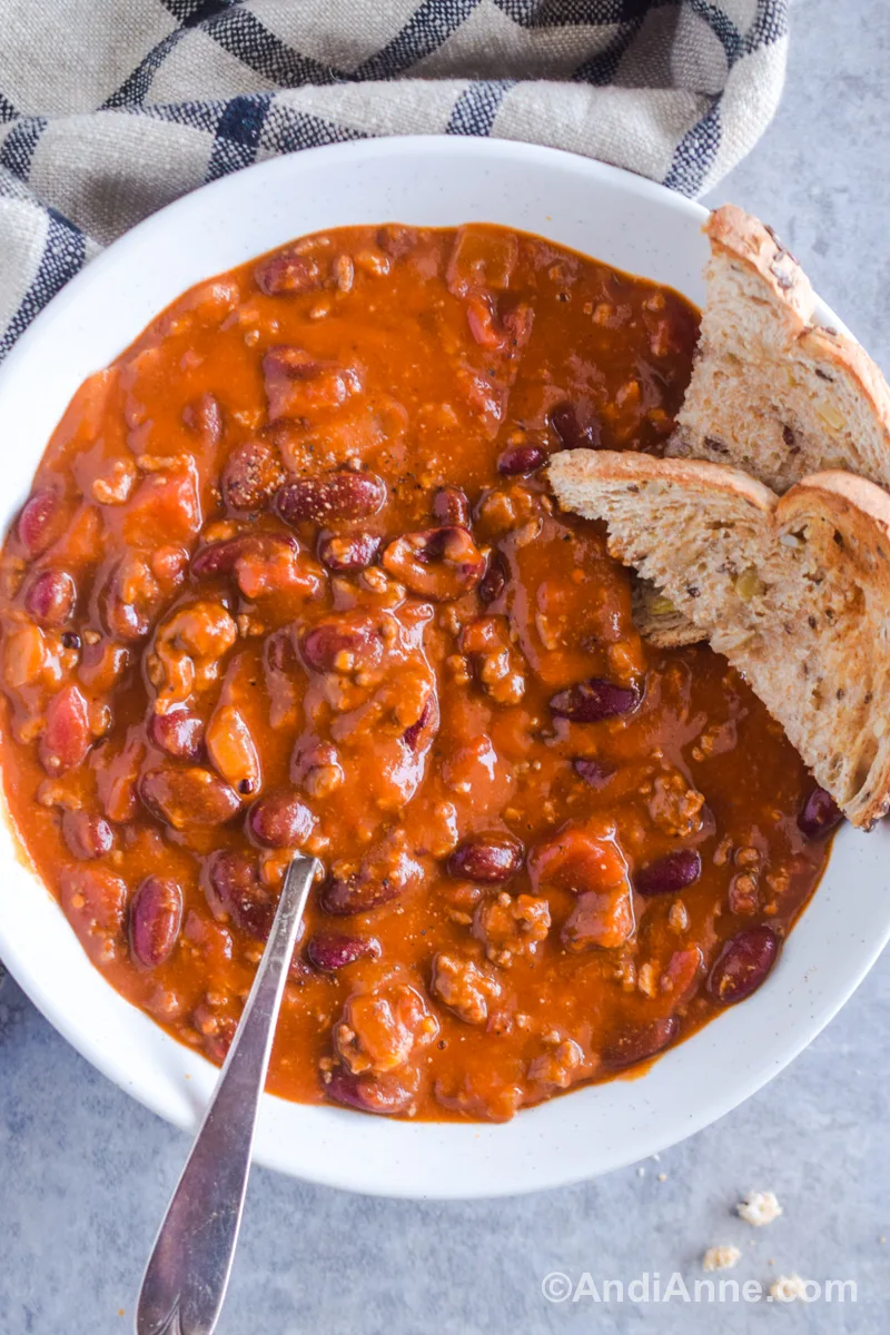 A white bowl with chili and a spoon. Slices of toast are on the side of the bowl.