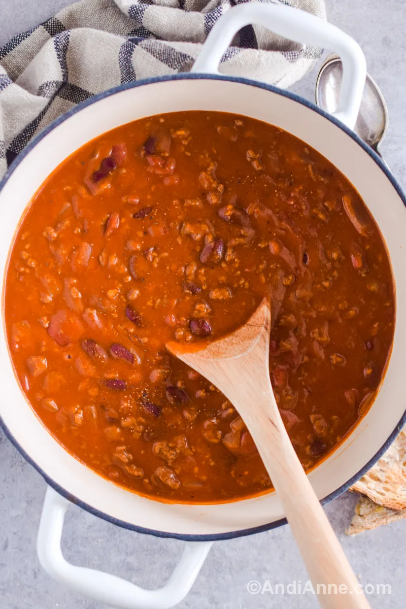 A white pot with chili inside and a wood spoon. Kitchen towel, metal spoons and sliced toast surround the pot.