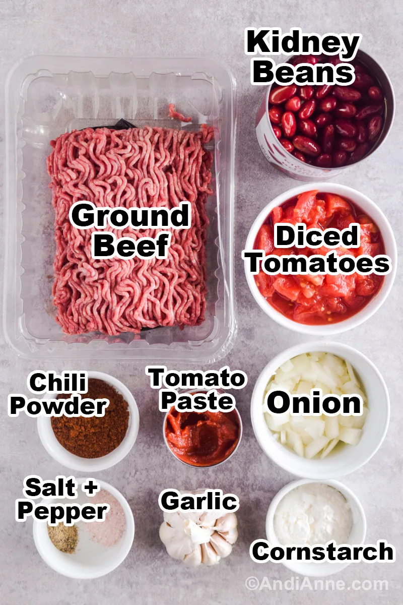 Recipe ingredients on the counter including raw ground beef, a can of kidney beans, and bowls of diced tomatoes, chili powder, tomato paste and chopped onion, cornstarch, salt and pepper and a garlic bulb.