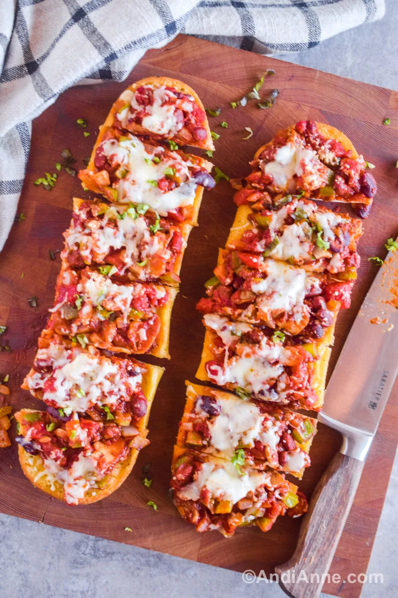 Sliced garlic bread loaves with chili and melted cheese and top. A knife beside them. All on a cutting board.