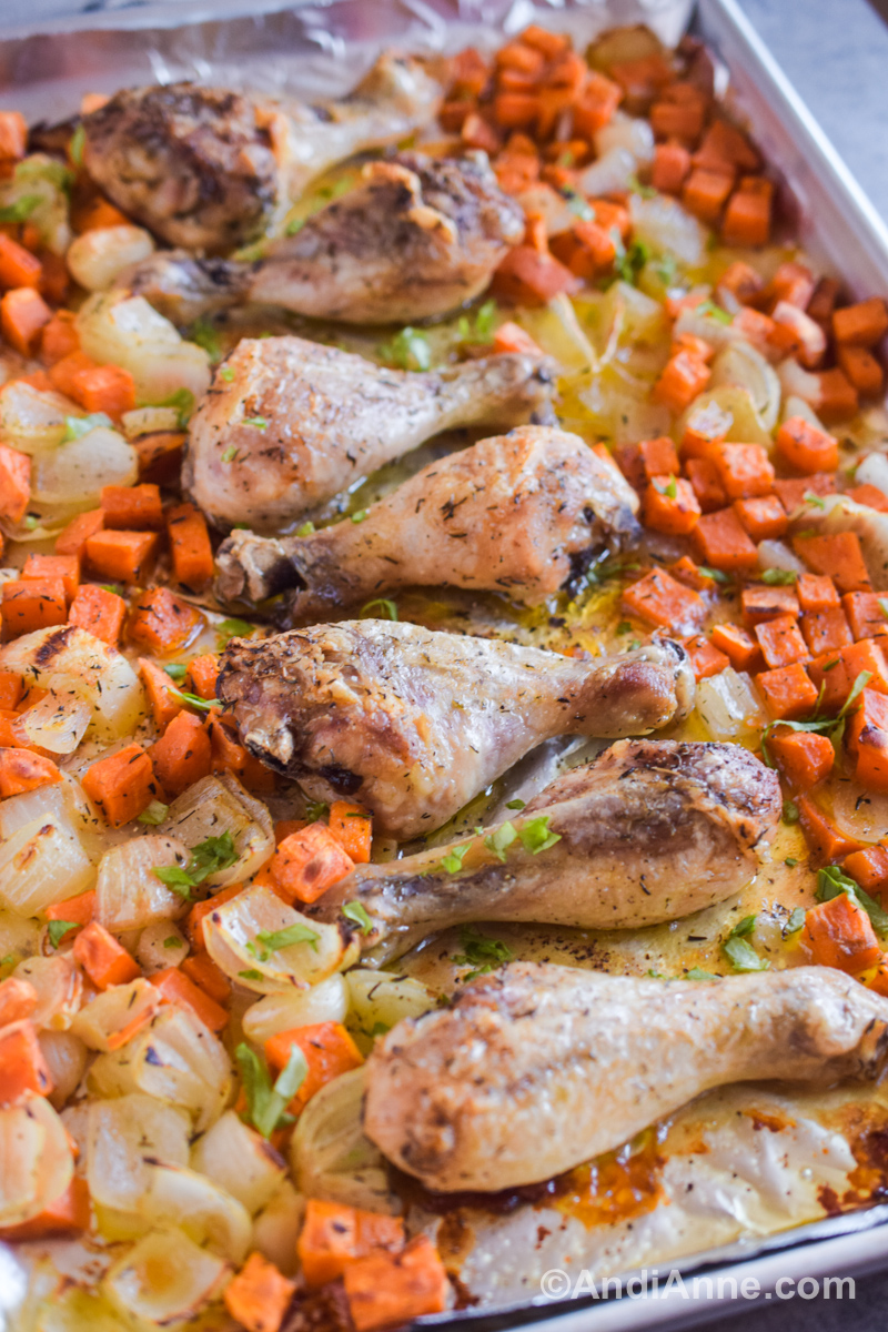 Close up of chicken drumsticks, chopped sweet potato, and onion on a baking sheet.
