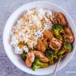 A white plate with cooked rice and chicken thighs with broccoli.