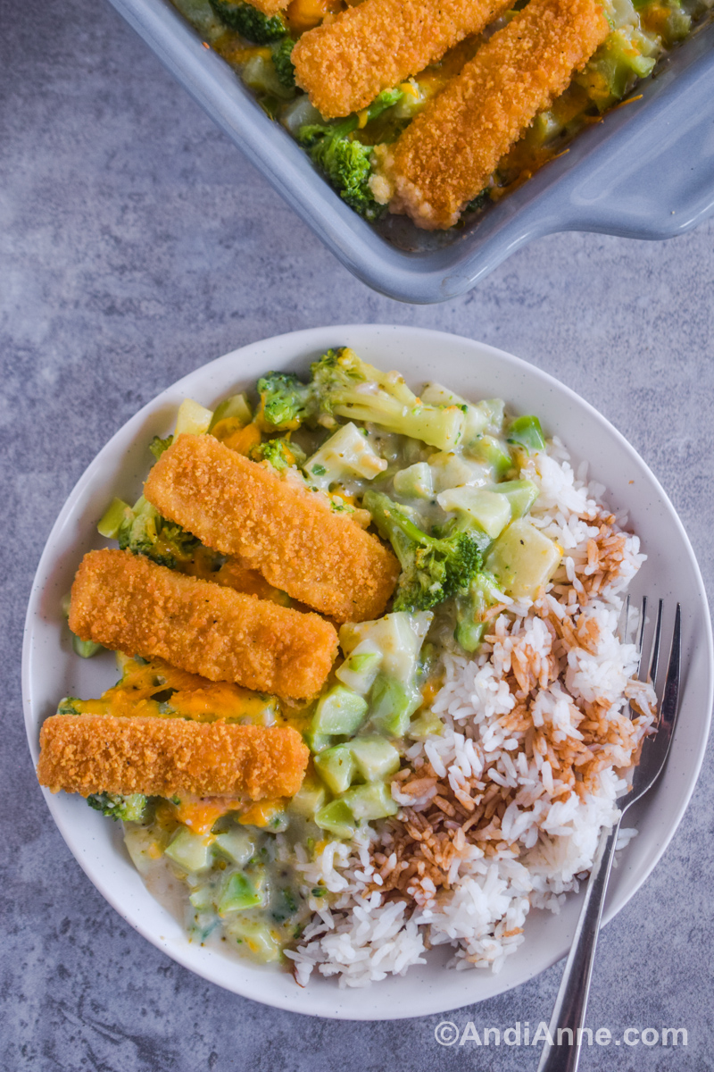 A white plate with three fish sticks, creamy chopped broccoli mixture and white rice with soy sauce on top.