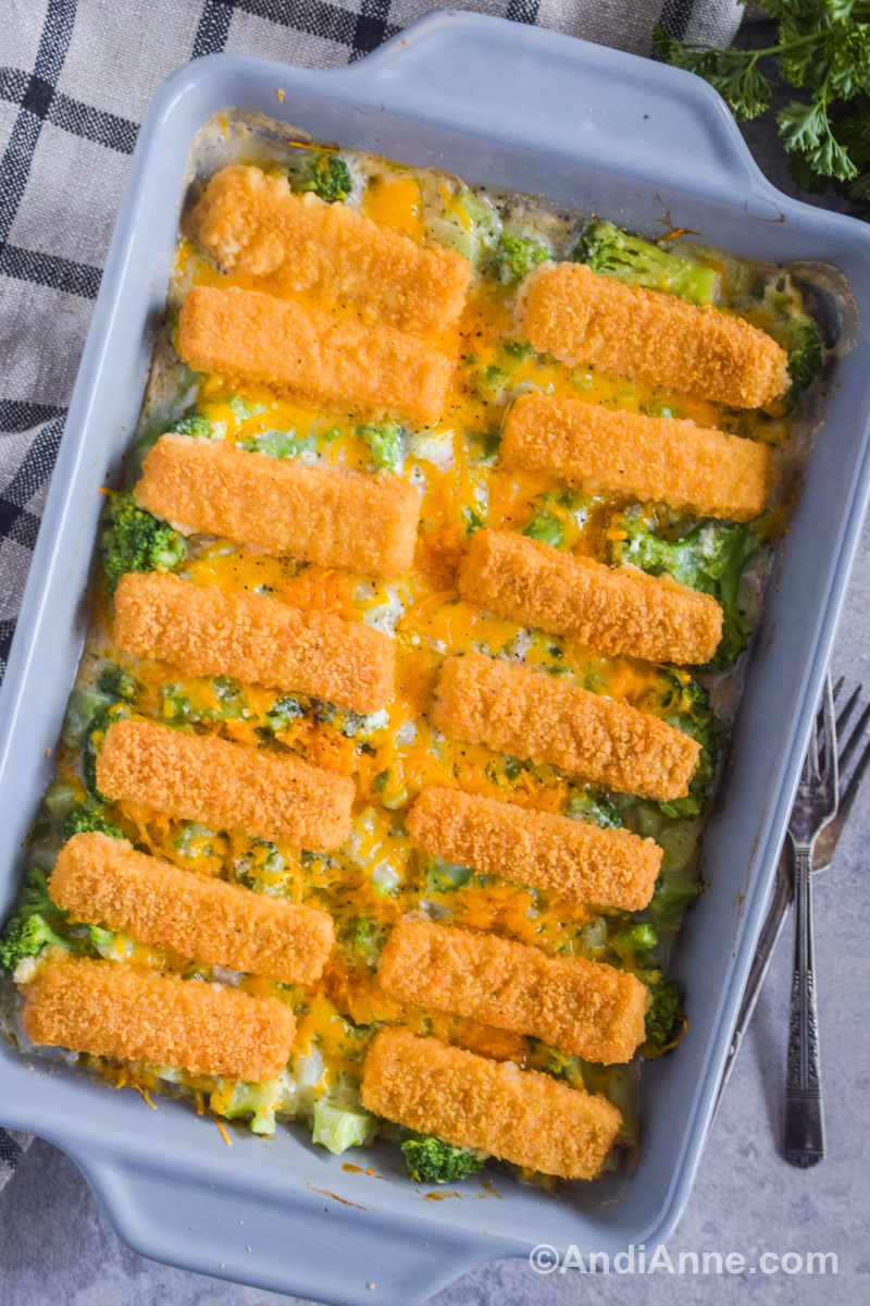 Close up of baked fish sticks on top of creamy broccoli mixture with shredded cheese. All in a grey cassrole dish. 