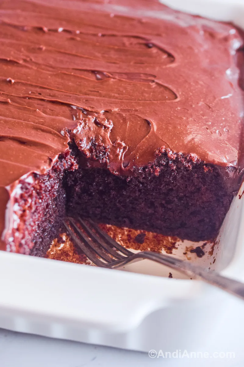 Close up of chocolate cake in a white dish with a fork and frosting on top.