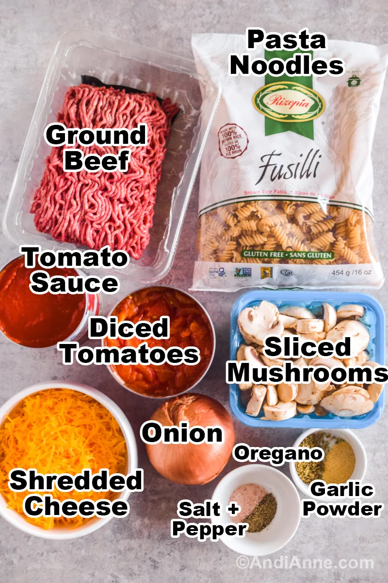 Ingredients to make the recipe on a counter including raw ground beef, a bag of pasta noodles, can of tomato sauce, diced tomatoes, sliced mushrooms, yellow onion, bowl of shredded cheddar cheese and bowls of spices.