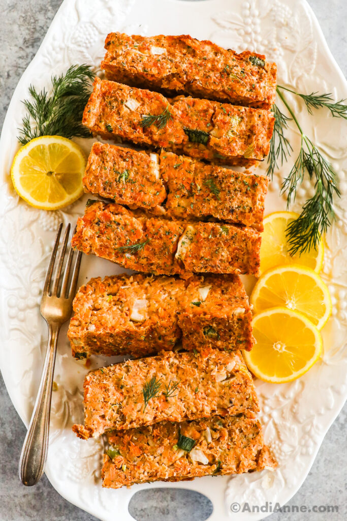 Slices of salmon loaf on a white plate with fresh dill, a fork and lemon slices.
