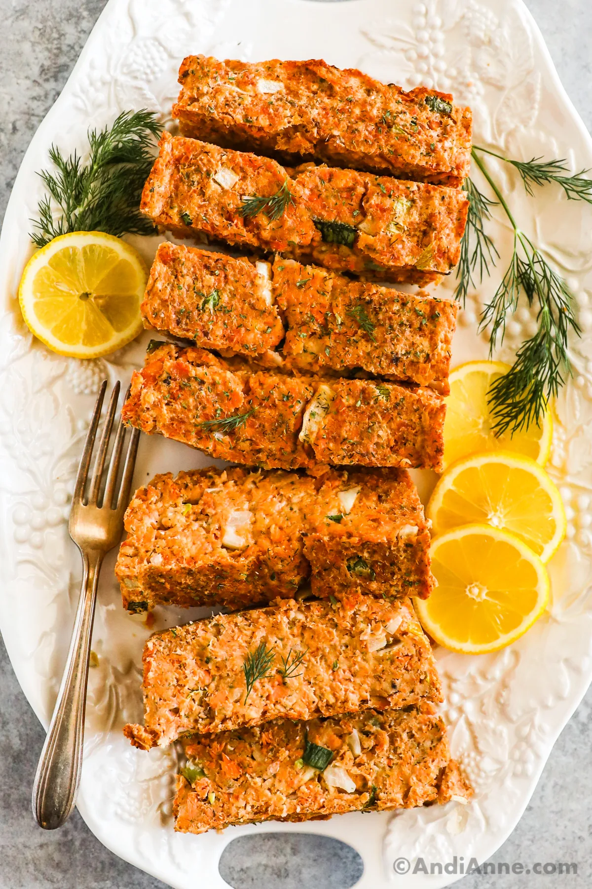 Slices of salmon loaf on a white plate with fresh dill, a fork and lemon slices.