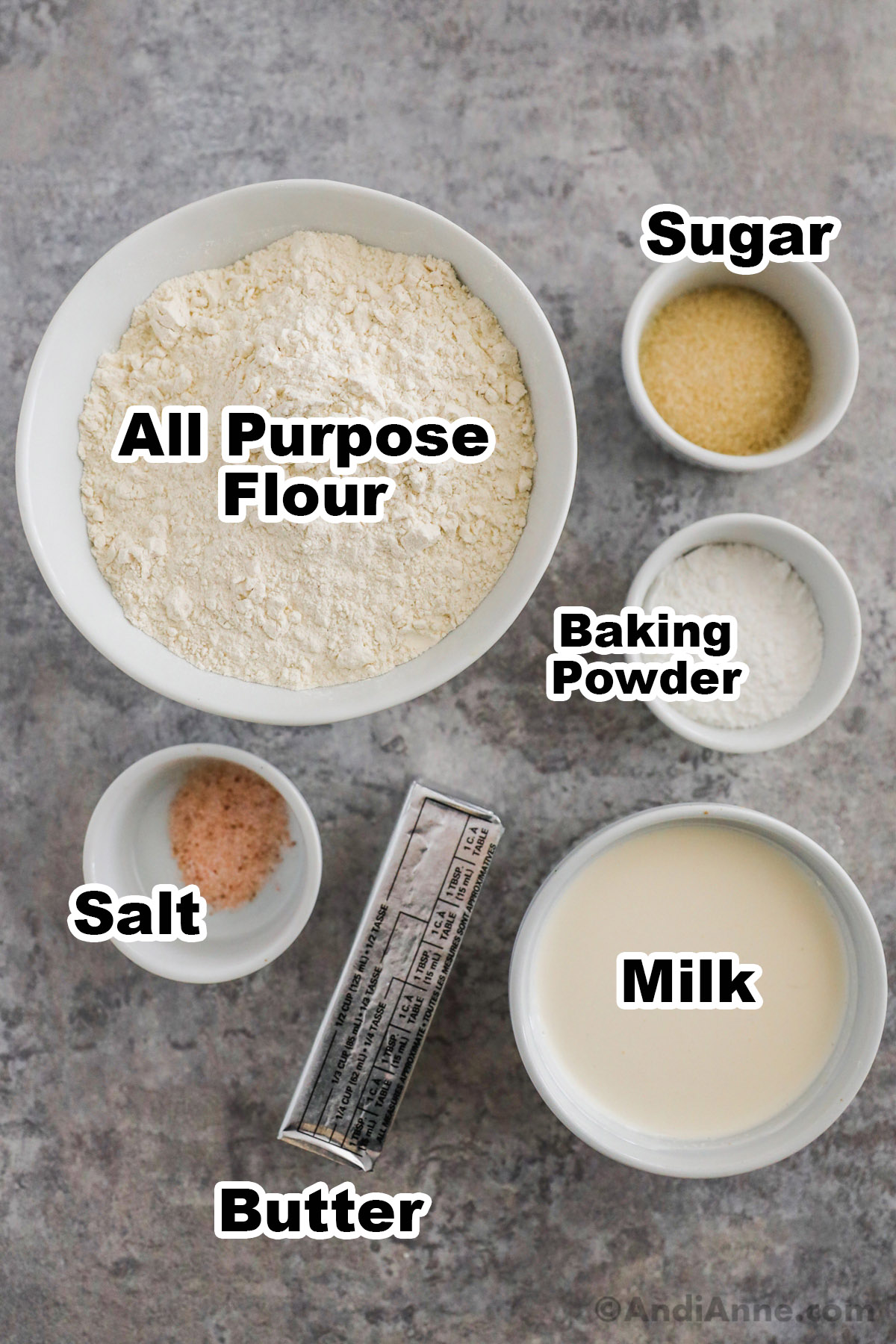 Recipe ingredients on counter including bowl of all purpose flour, bowls of sugar, baking powder, salt, milk and a stick of butter.