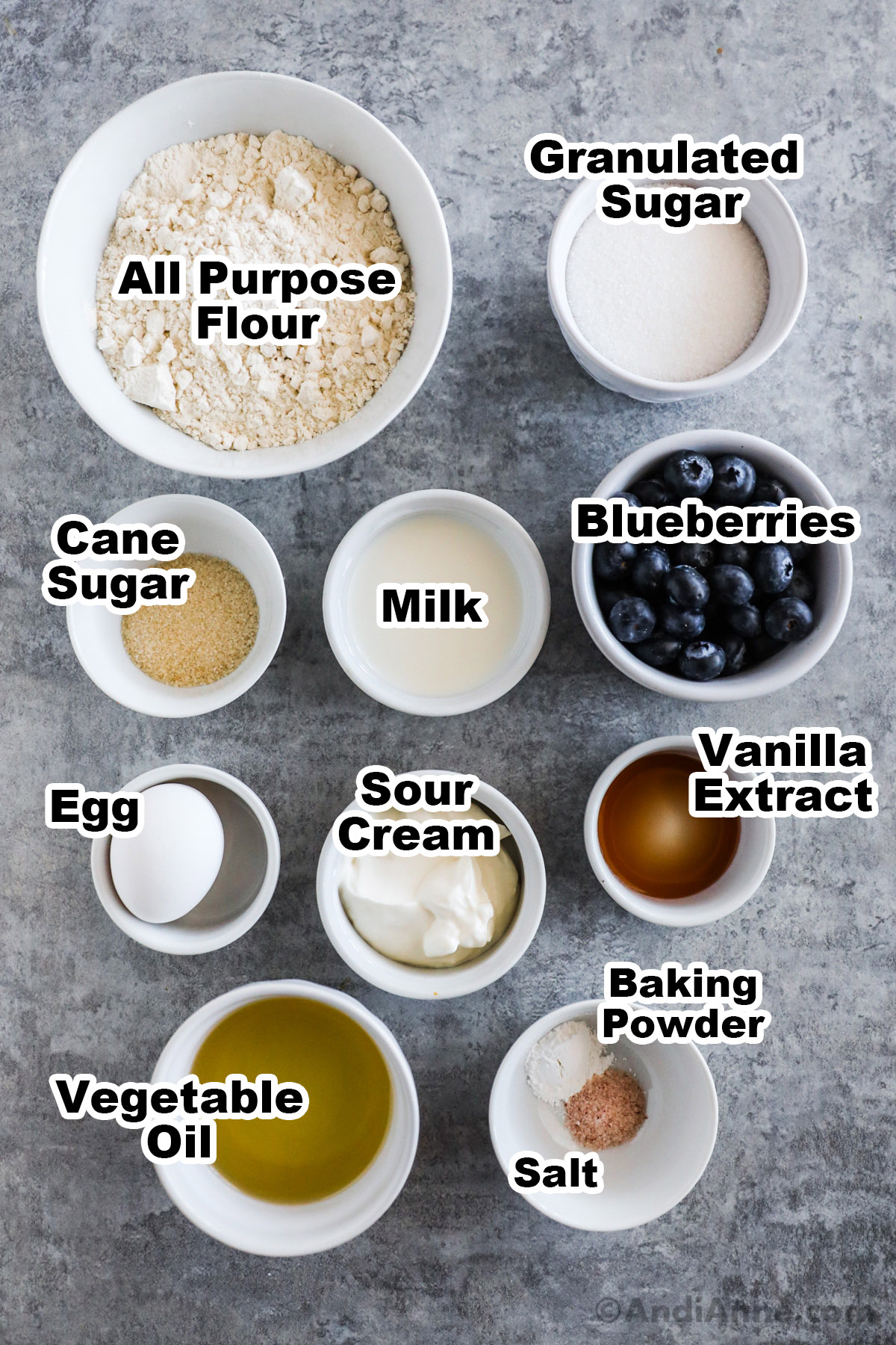 Recipe ingredients in bowls including flour, sugar, milk, blueberries, egg, sour cream, vanilla extract, oil, and baking powder.
