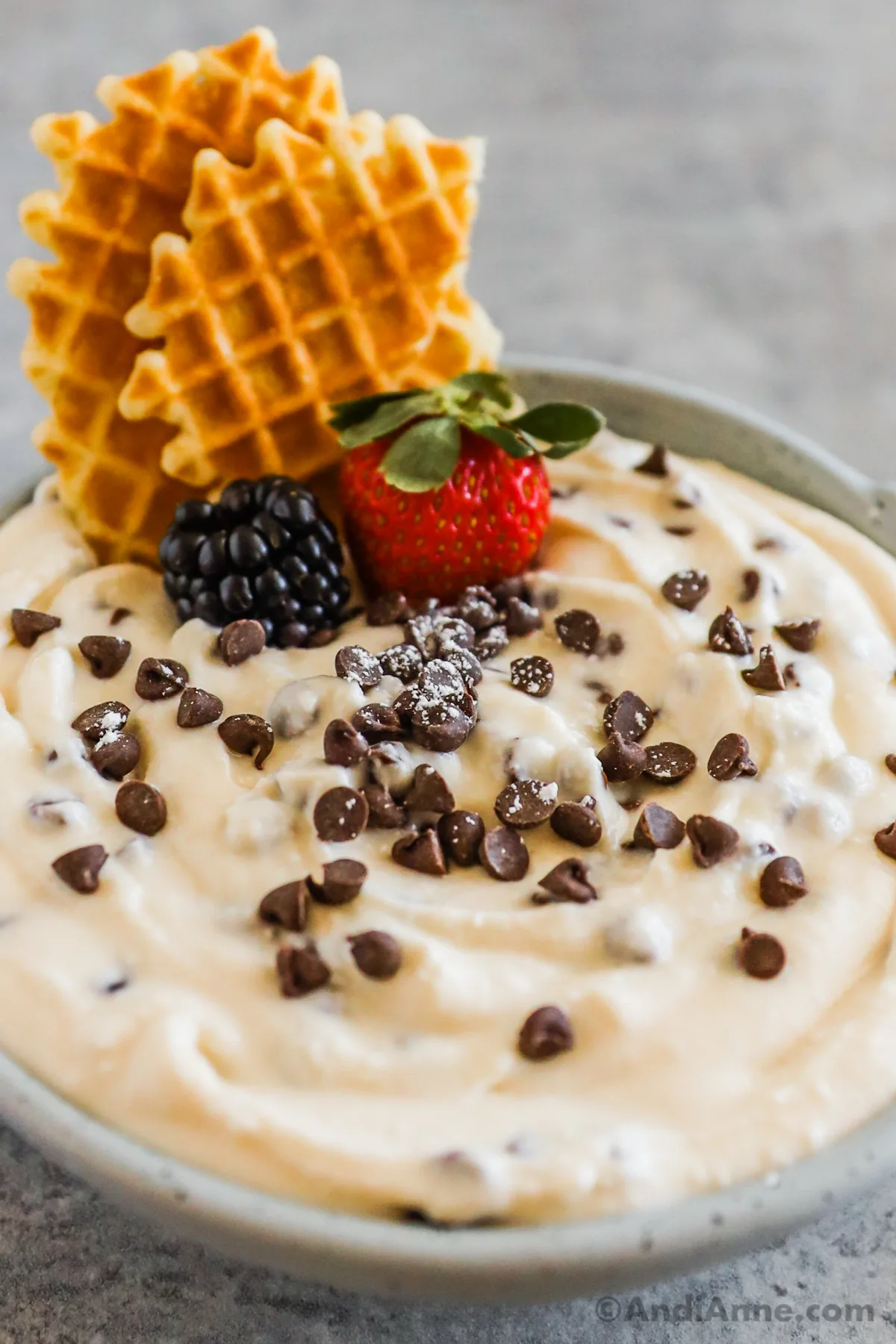 A bowl of cannoli dip recipe topped with mini chocolate chips, a strawberry, blackberry, and two waffle cookies.