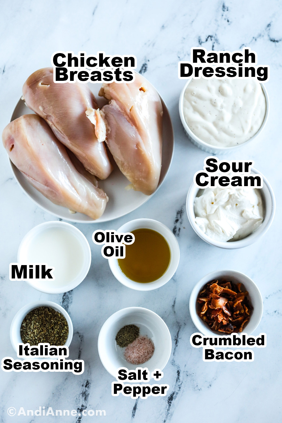 Recipe ingredients on the counter including raw chicken breasts, bowls of ranch dressing, sour cream, milk, olive oil, crumbled bacon, italian seasoning, salt and pepper.