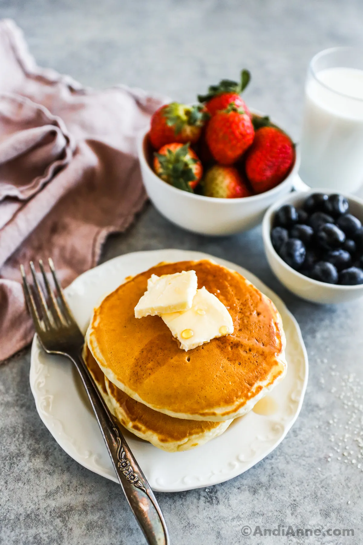 Pancakes on a white plate with butter and a fork. A bowl with strawberries, bowl with blueberries, glass of milk and napkin behind it. 