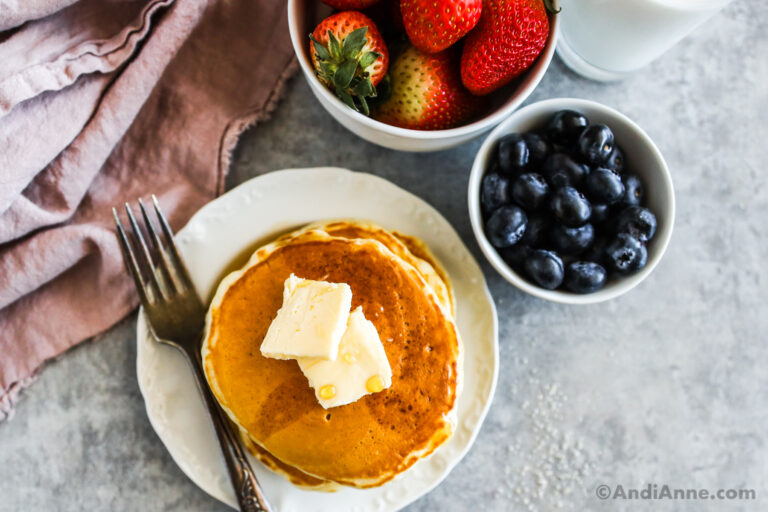 A white plate of pancakes topped with syrup and butter and with bowls of blueberries and strawberries.