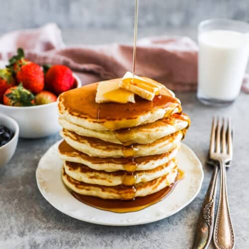 Stack of fluffy homemade pancakes with butter and maple syrup and bowls of strawberries and glass of milk in background.