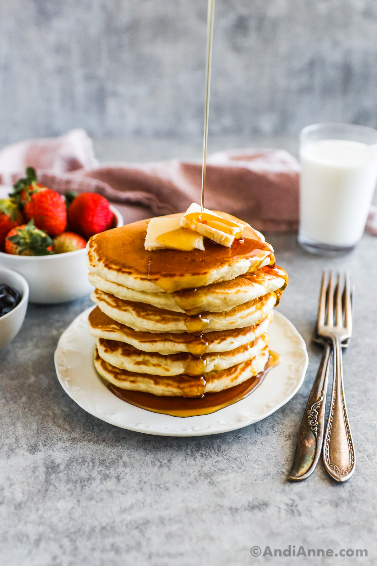 A stack of fluffy pancakes on a white plate with butter and maple syrup. Forks, a glass of milk, a bowl of strawberries in the background.