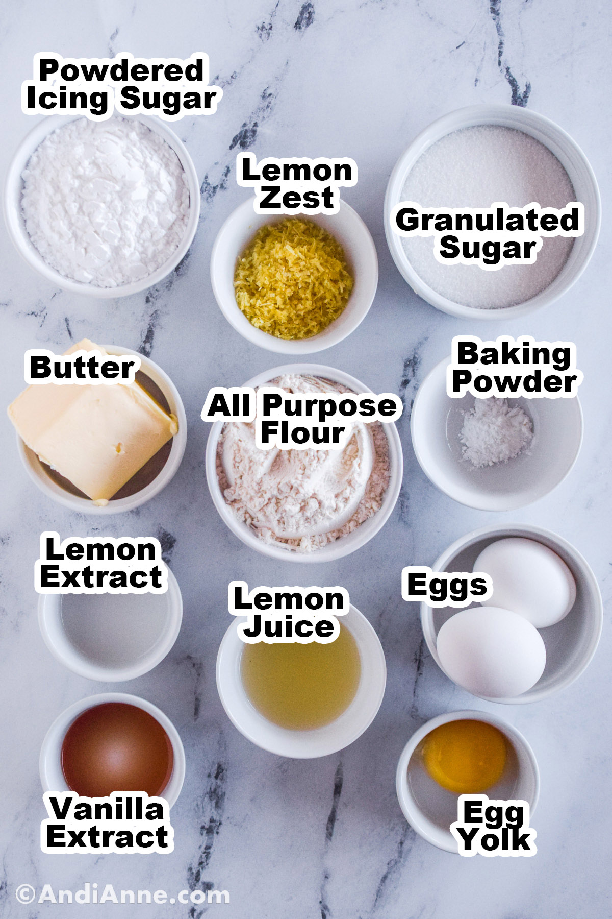 Recipe ingredients on the counter in bowls including powdered icing sugar, lemon zest, granulated sugar, butter, flour, baking powder, lemon extract, lemon juice, eggs, vanilla extract and egg yolk.