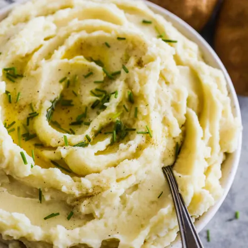 Close up of mashed potatoes with chives on top and a spoon. Russet potatoes in the background.