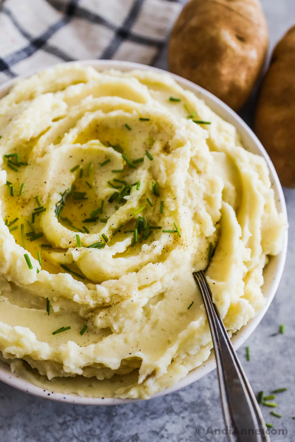 Close up of mashed potatoes with chives on top and a spoon. Russet potatoes in the background.