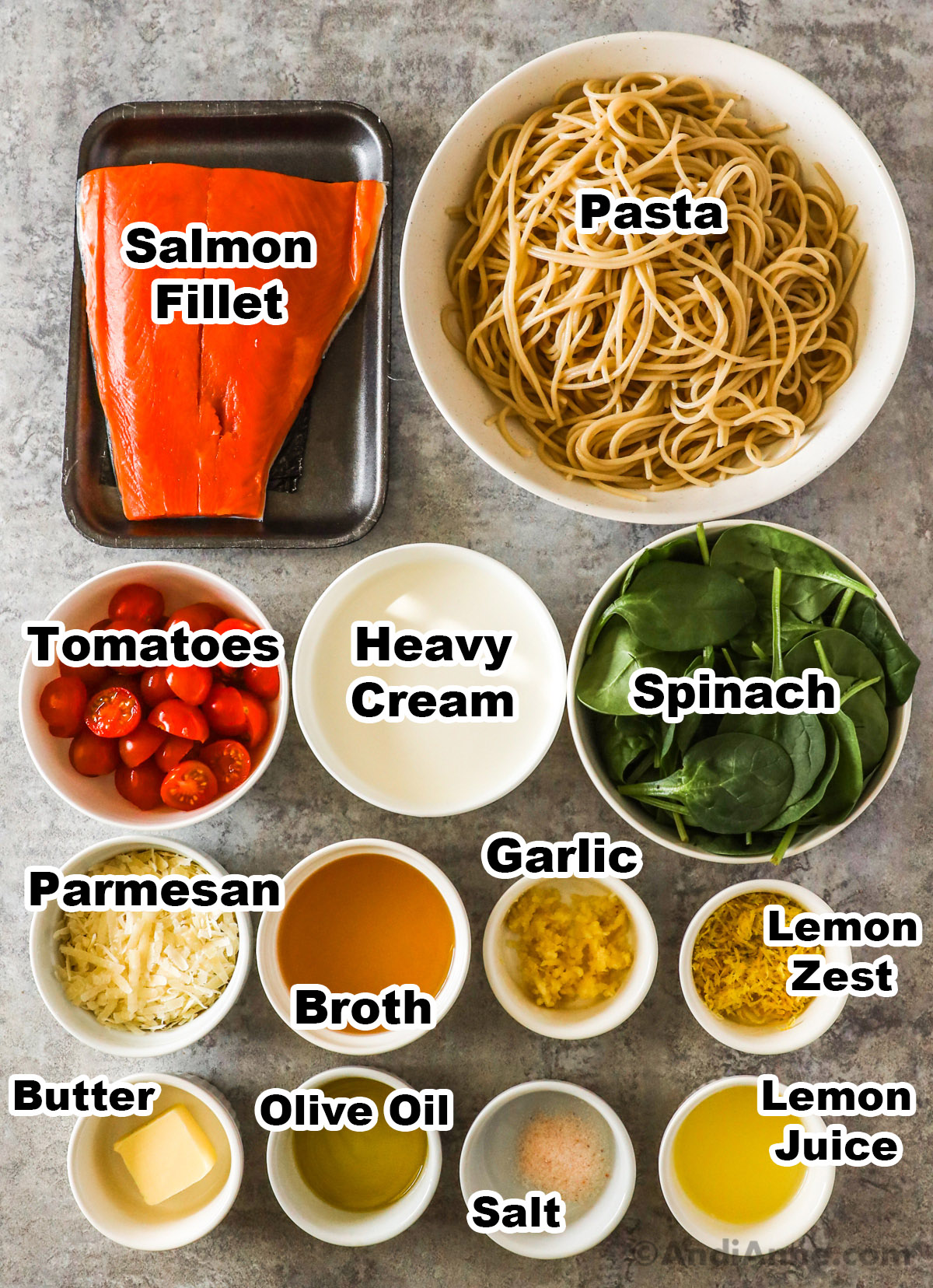 Recipe ingredients including a salmon fillet, bowl of cooked pasta, sliced cherry tomatoes, heavy cream, spinach, grated parmesan, broth, garlic, lemon zest, butter, olive oil, salt and lemon juice.