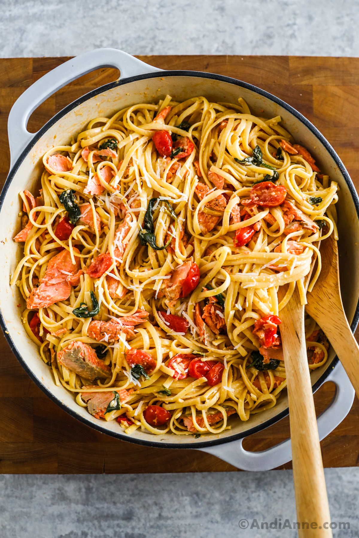 A large pot with salmon pasta recipe inside with tomatoes and spinach and two wood spoons. 