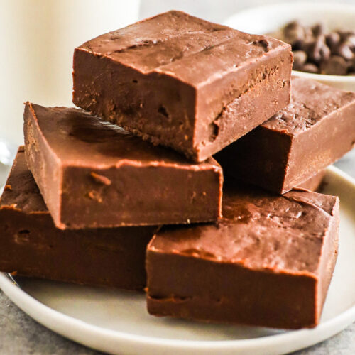 A stack of chocolate fudge on a plate with glass of milk in the background.
