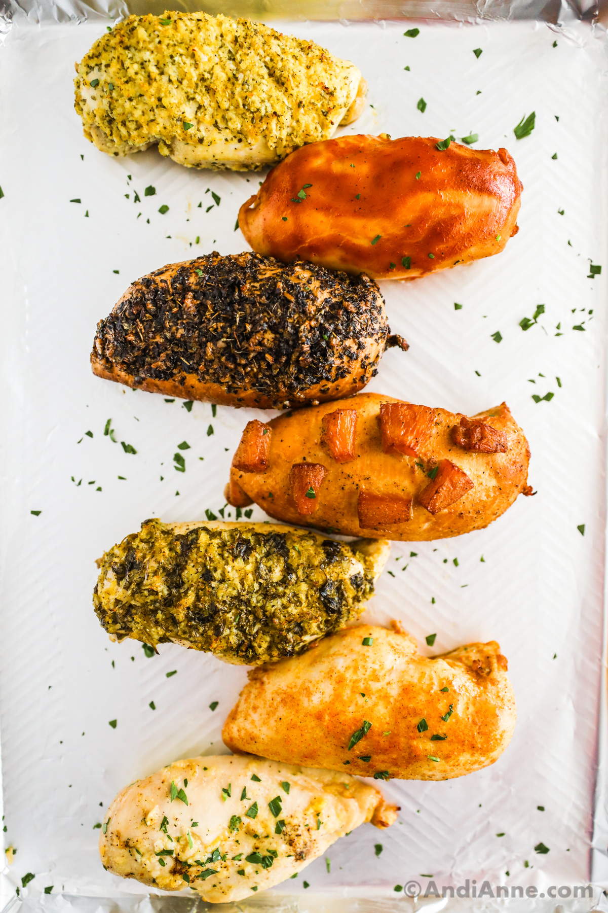 8 baked chicken breasts with various seasonings on top of each all sitting on a baking sheet.