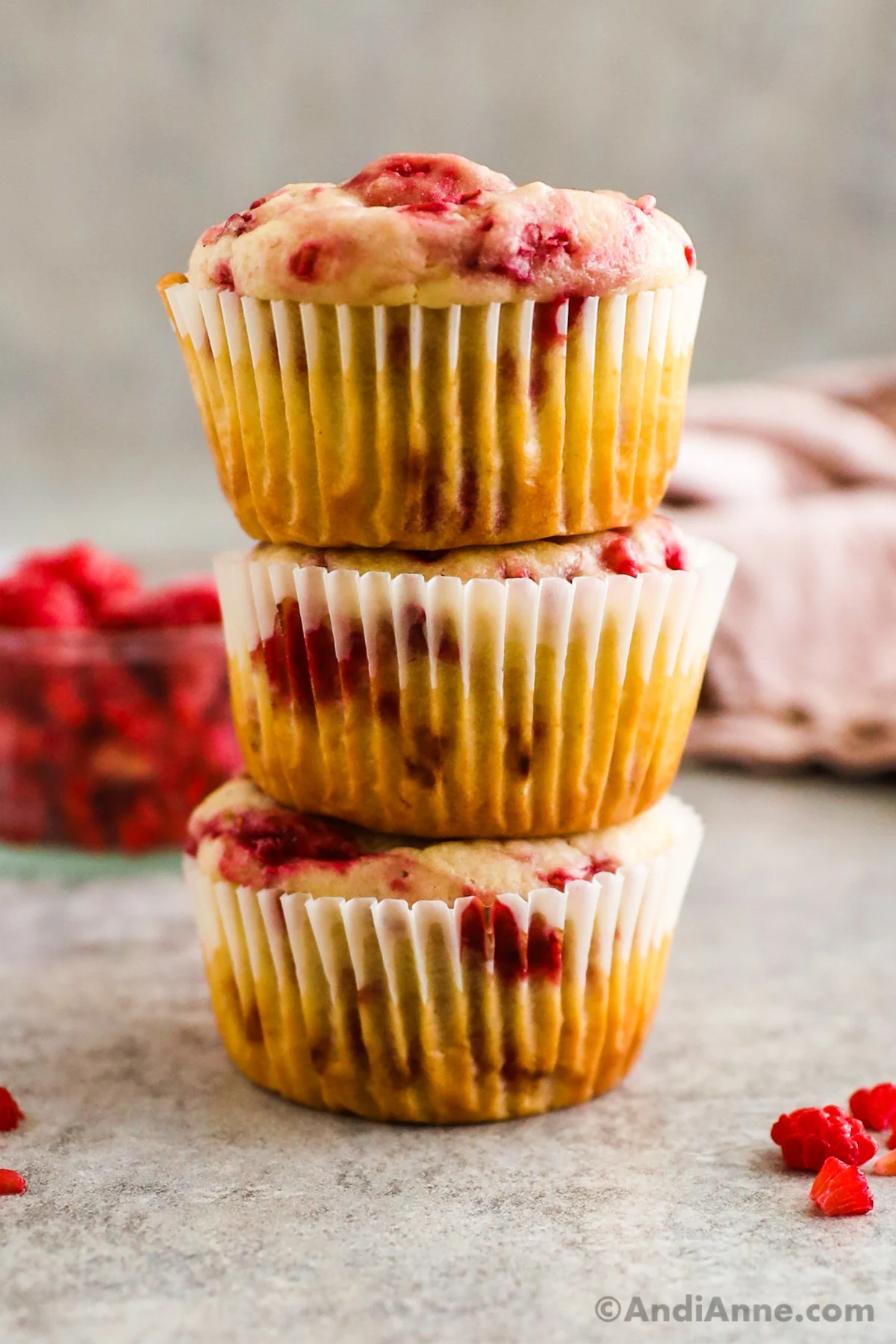 Three muffins stacked on top of each other with a bowl of raspberries behind it. 