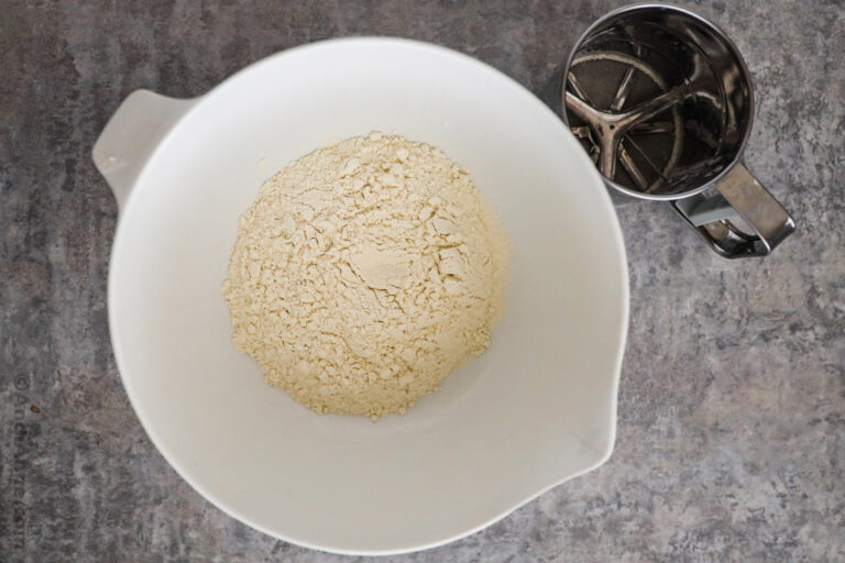 A white bowl with dry flour ingredients and a sifter beside.