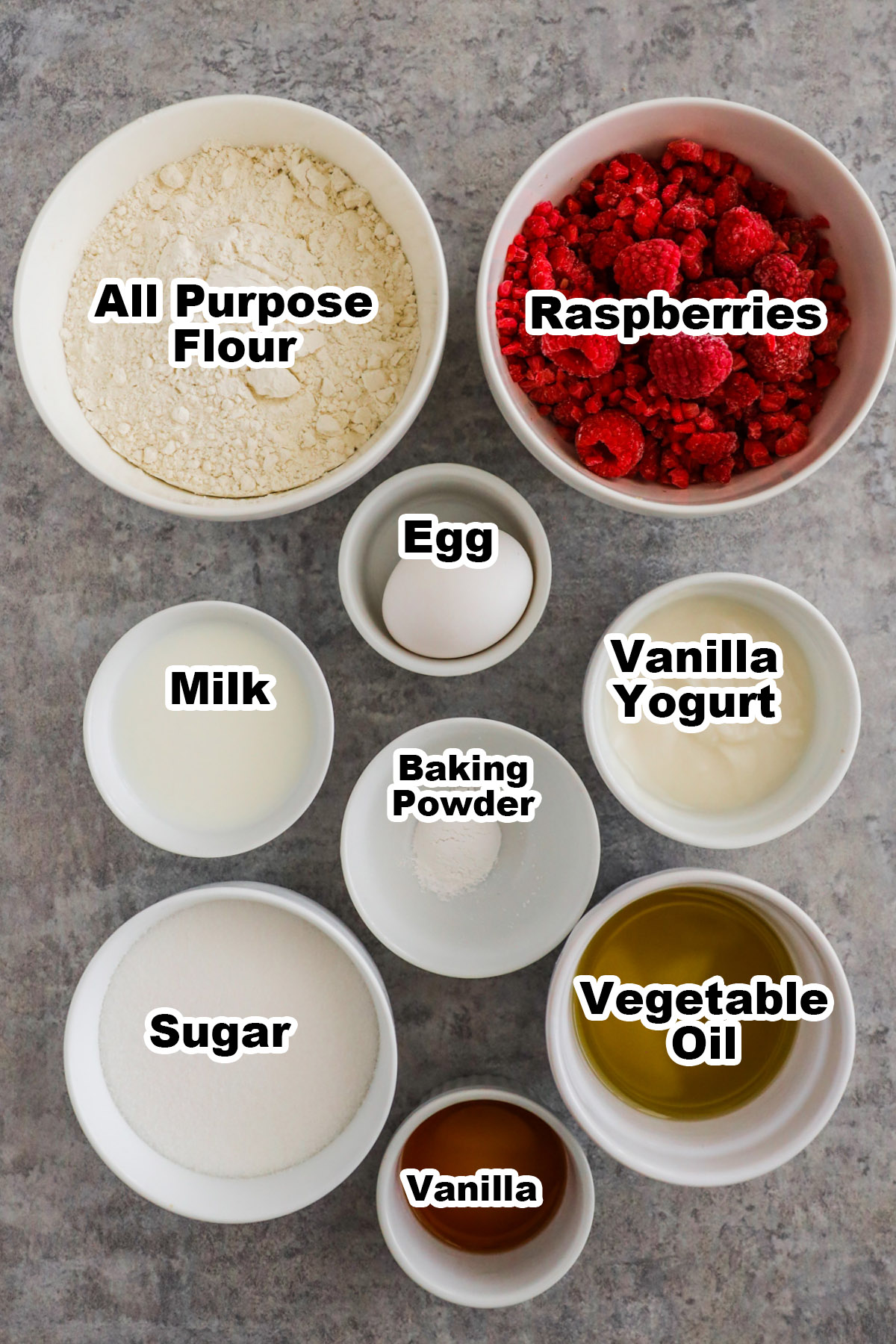 Recipe ingredients on the counter in bowls including flour, frozen raspberries, egg, vanilla yogurt, milk, baking powder, vegetable oil, sugar and vanilla extract.