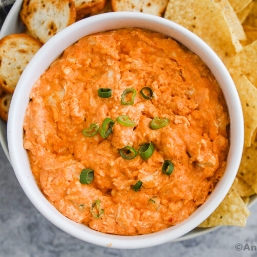 a white bowl of buffalo chicken dip with tortilla chips and crackers on a plate.