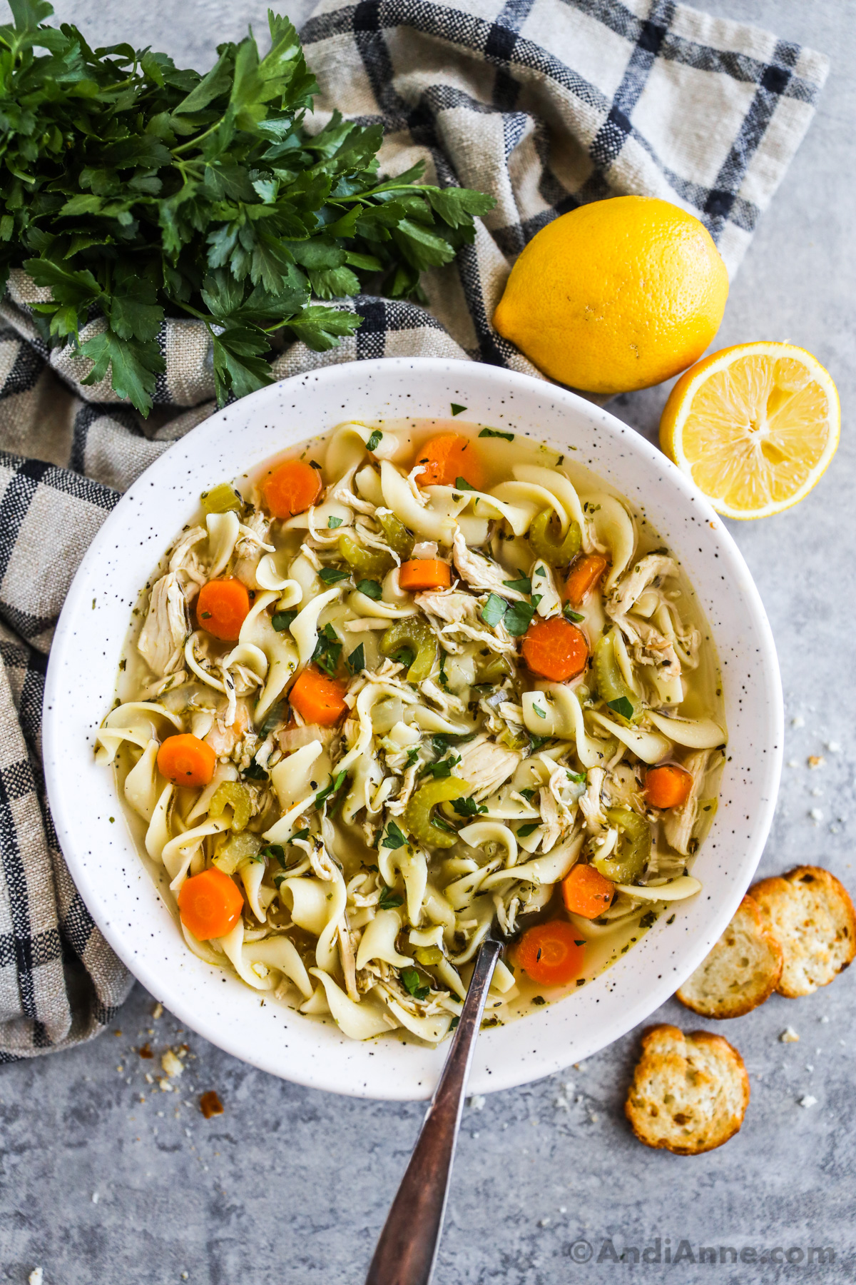 a bowl of chicken noodle soup surrounded by lemon, parsley and a kitchen towel.