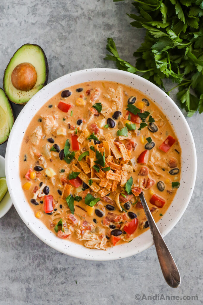 A bowl of chicken taco soup with spoon. Sliced avocado and parsley are around the bowl.